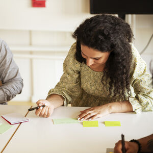 Kaksi henkilöä istuu pöydän ääressä tarralappujen, kynien ja muistikirjan kanssa ja tekevät yhteistyötä.
