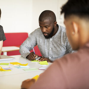 Kolme ihmistä tekee yhteistyötä pöydän ääressä, jossa on muistilappuja ja kannettavia tietokoneita.