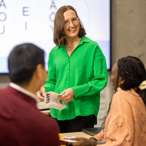 Opettaja vihreässä paidassa ohjaa kahta oppilasta luokkahuoneessa, jossa on valkotaulu, jossa näkyy vokaaleja.