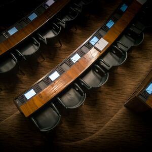 An overhead view of a legislative chamber with curved desks and black chairs, each desk equipped with blue screens.