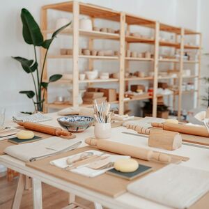 Trähyllor med keramik bakom ett bord med konstmaterial. En stor växt syns i hörnet.