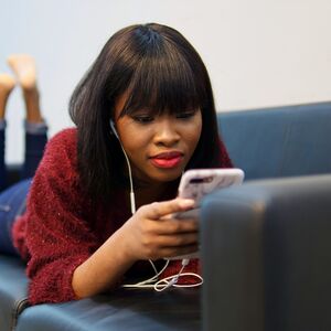 Person lying on a black sofa, using a smartphone with earphones in. They are wearing a red sweater and blue jeans.