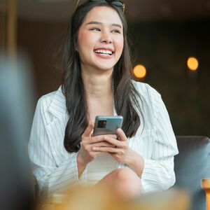 En person med långt hår sitter inomhus och använder en smartphone. Hen har på sig en ljusrandig skjorta.