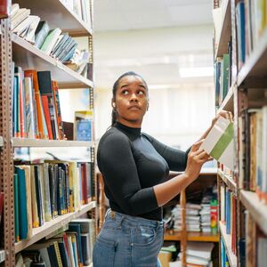 Person standing between shelves in a library, holding a book. Shelves are filled with books.
