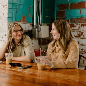 Kaksi naista istuu puupöydän ääressä, heidän edessään on jääjuomia. Vasemmalla on ikkuna ja ruukkukasvi.