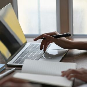 Personer arbetar på bärbara datorer med anteckningsböcker och överstrykningspennor på ett träbord nära fönster.