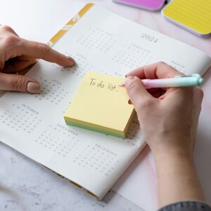 Händer skriver en att-göra-lista på en klisterlapp på en kalender från 2024, med anteckningsböcker och gem i närheten.