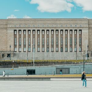 Riksdagshuset. En stor stenbyggnad med kolonner och rader av fönster. Människor går framför och träd flankar sidorna.
