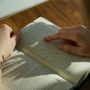 Närbild av händer som läser en öppen bok på ett träbord, med ett finger som pekar på en textrad.