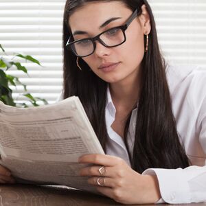 En person med långt mörkt hår läser en tidning vid ett träbord inomhus. Det finns växter och persienner i bakgrunden.