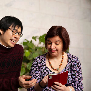 Kaksi henkilöä katsovat punaista puhelinta. Toinen henkilö on pukeutunut viininpunaiseen neuleeseen ja toinen kukkapaitaan ja helmikoruun.