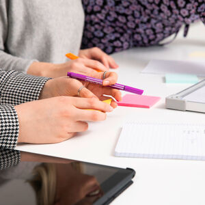 Ihmiset työskentelevät pöydän ääressä muistikirjojen, muistilappujen ja tabletin kanssa. Yksi pitelee violettia kynää.