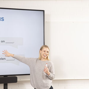 Henkilö osoittaa suurta näyttöä, jossa on teksti 'Harjoitus', ja pitelee tussia. Toinen henkilö etualalla.