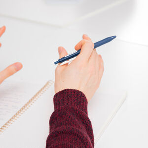 Henkiö viininpunaisessa villapaidassa pitää kynää ja elehtii muistion äärellä valkoisella pöydällä.