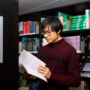 Person i röd tröja läser en bok på ett bibliotek, står mellan höga bokhyllor fyllda med olika böcker.