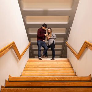 Two people standing at the top of a wooden staircase, pointing at a tablet. The stairs have wooden handrails.