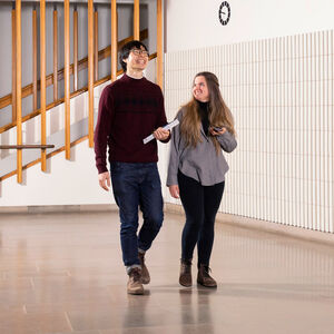 Kaksi henkilöä kävelee sisätiloissa. Miehellä on muistiinpanovihko ja punainen neule. Naisella on puhelin ja harmaa paita.