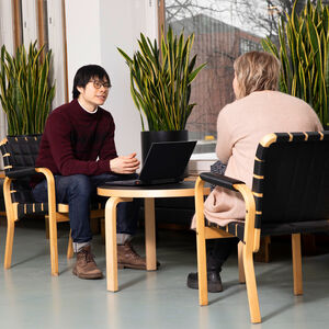 Kaksi ihmistä istuu pienen pöydän ääressä modernissa huoneessa, jossa on isot ikkunat, kasveja ja puuhuonekaluja. Toisella on kannettava tietokone auki.
