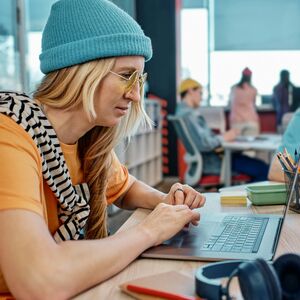 Kaksi henkilöä työskentelee pöydällä, jossa on kannettavia ja muistikirjoja modernissa toimistossa. Taustalla keskustellaan.