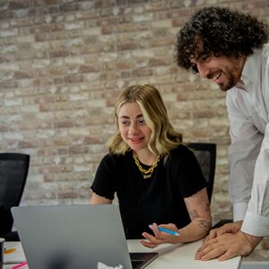 Kaksi ihmistä toimistossa, yksi istuu kannettavan ääressä näyttäen jotain seisovalle ja nojaavalle henkilölle.