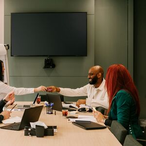 Neljä ihmistä kokouksessa neuvotteluhuoneessa, jossa on iso näyttö seinällä. Kaksi henkilöä vaihtaa tavaraa.