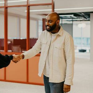 Kaksi henkilöä kättelee toimistossa, taustalla on punaiset väliseinät ja työpöytiä.