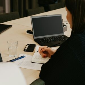 Henkilö kirjoittaa muistikirjaan pöydällä, jossa on kannettava tietokone, vesilasi, tabletti ja kynä.