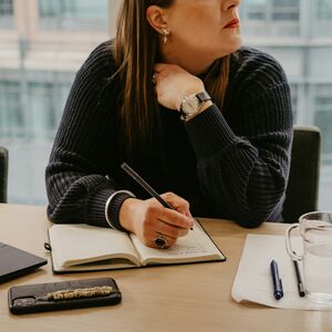 A person writes in a notebook at a desk with a laptop, phone, pen, paper, and glass of water.