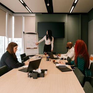 Henkilö esittelee valkotaululla kokoushuoneessa, jossa kolme osanottajaa istuu pöydän ympärillä kannettavien tietokoneiden kanssa.