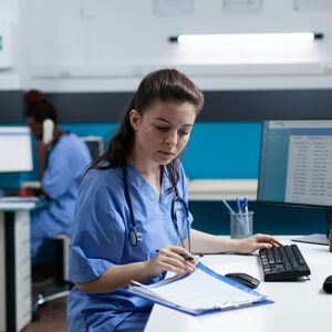 Kaksi terveydenhuollon ammattilaista työskentelee tietokoneilla, toinen kirjoittaa ja toinen puhuu puhelimeen.