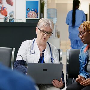 Lääkäri keskustelee potilaan kanssa odotustilassa kannettavan tietokoneen äärellä, taustalla on toinen potilas ja hoitaja.