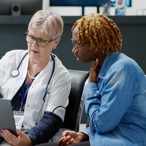 Lääkäri ja henkilö istuvat tuoleilla odotustilassa ja katsovat kannettavaa tietokonetta. Lääkärillä on stetoskooppi.