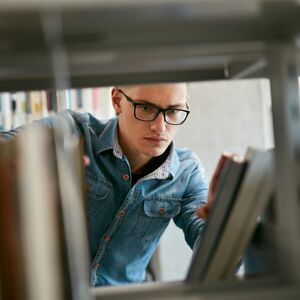 Kirjasto, jossa henkilö ulottuu kirjaan, päällään farkkupaita, kirjahyllyjen välissä.