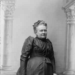 Nainen tummassa viktoriaanisessa mekossa seisoo kahden koristeellisen pylvään välissä, pitäen oikeassa kädessään laukkua.