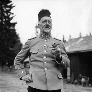 Ilmari Kianto sotilaspuvussa seisoo ulkona pitäen savuketta, toinen käsi lantiolla. Taustalla puita ja rakennus.