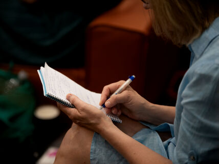 Henkilö kirjoittaa muistiinpanoja. Hänellä on kädessä muistivihko ja kynä.