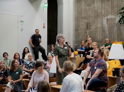 Ryhmä ihmisiä istuu ja kuuntelee keskellä seisovaa naista jolla on mikrofoni kädessä.