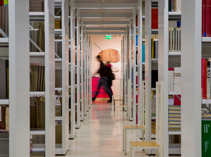 Krjaston hyllyjä joiden takana kulkee ihmisiä.