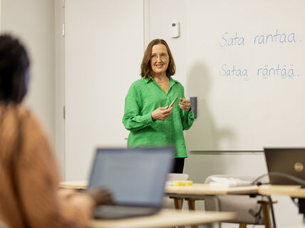 Opettaja seisoo luokan edessä. Taululla lukee "Sata rantaa. Sataa räntää."