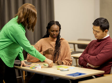 Opettaja ja kaksi opiskelijaa katsovat pöydällä olevaa kortipeliä.