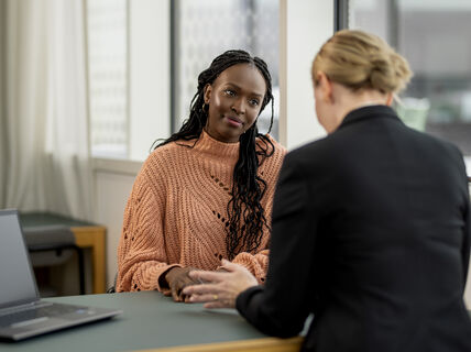 Two people having a conversation in an office setting, one wearing a peach sweater, the other in a black jacket. A laptop is on the table.
