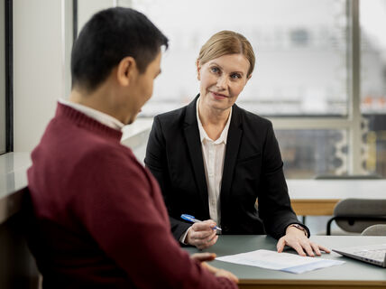 Opiskelija hymyilee ja keskustelee opettajan kanssa pöydän ääressä. Pöydällä on tietokone. Opiskelija katsoo hymyilevä opettajaa, jolla on kynä kädessä. Pöydällä on paperi ja tietokone.