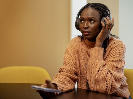 Opiskelija jolla on puhelin kädessä ja kuulokkeet päässä istuu pöydän ääressä.