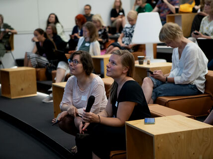 Ihmisiä istuu yleisössä. Eturivissä istuvalla naisella on mikrofoni kädessä.