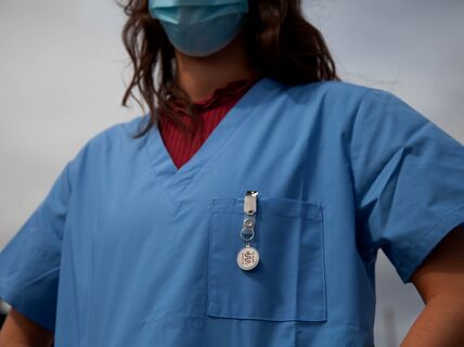 A person in blue scrubs and a blue face mask with a badge clipped to the pocket, standing against a cloudy sky.