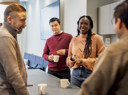 Ihmisiä keskustelemassa pöydän ääressä kahvimukien kanssa toimistoasetelmassa.