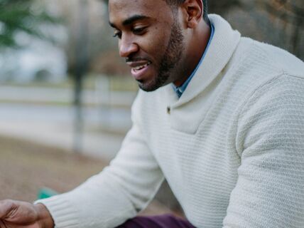 En person i vit tröja sitter på en parkbänk och läser en bok med röd och vit omslag.