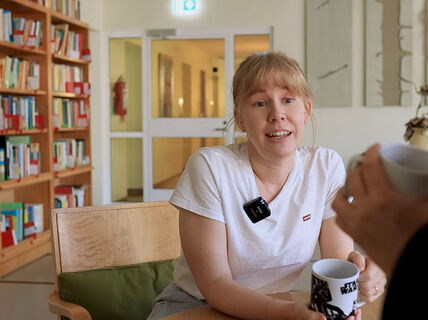 Two people having a conversation at a table, holding mugs. Bookshelves and a hallway are visible in the background.