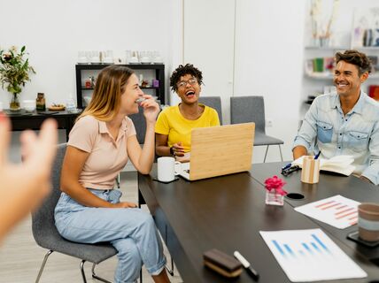 Three people at a meeting table with a laptop and documents. A person is gesturing in the foreground.