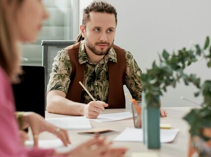 Ryhmä ihmisiä kokouksessa pöydän ääressä papereiden ja kynien kanssa. Yhdellä on vihreä kuviollinen paita ja ruskea liivi.